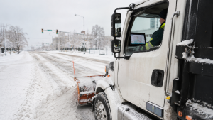 Snow plow truck clearing a snow-covered street, driver inside the cab with a map on the screen visible through the window